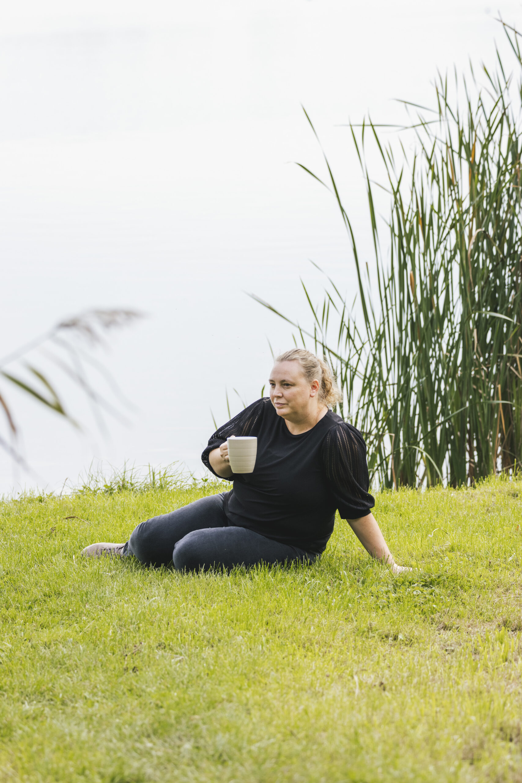 Lindsy zit rustig in het gras aan het water met een kopje in de hand en neemt een moment om stil te staan bij wat ze voelt
